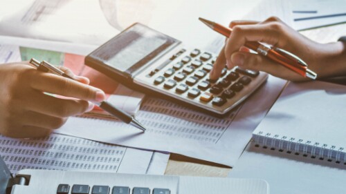 two professionals crunching numbers with pens and a calculator amidst paper documents, a journal, and a laptop