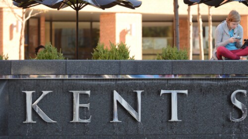 student looking at phone and sitting on sign on campus that says Kent State