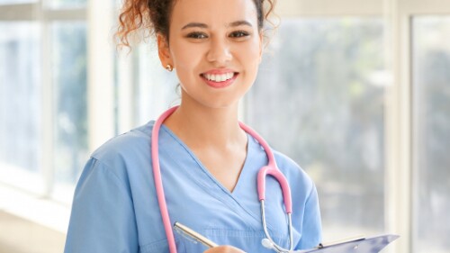 nurse wearing a stethoscope and holding a clipboard smiles in front of a window in a clinic