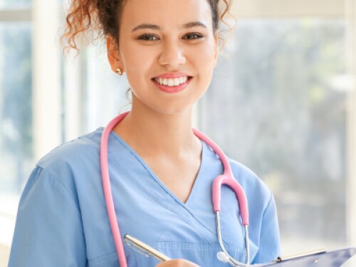nurse wearing a stethoscope and holding a clipboard smiles in front of a window in a clinic