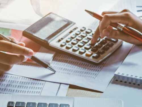 two professionals crunching numbers with pens and a calculator amidst paper documents, a journal, and a laptop