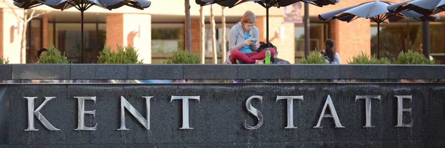 student looking at phone and sitting on sign on campus that says Kent State