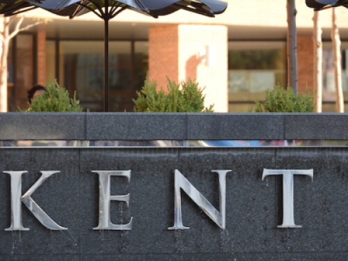 student looking at phone and sitting on sign on campus that says Kent State